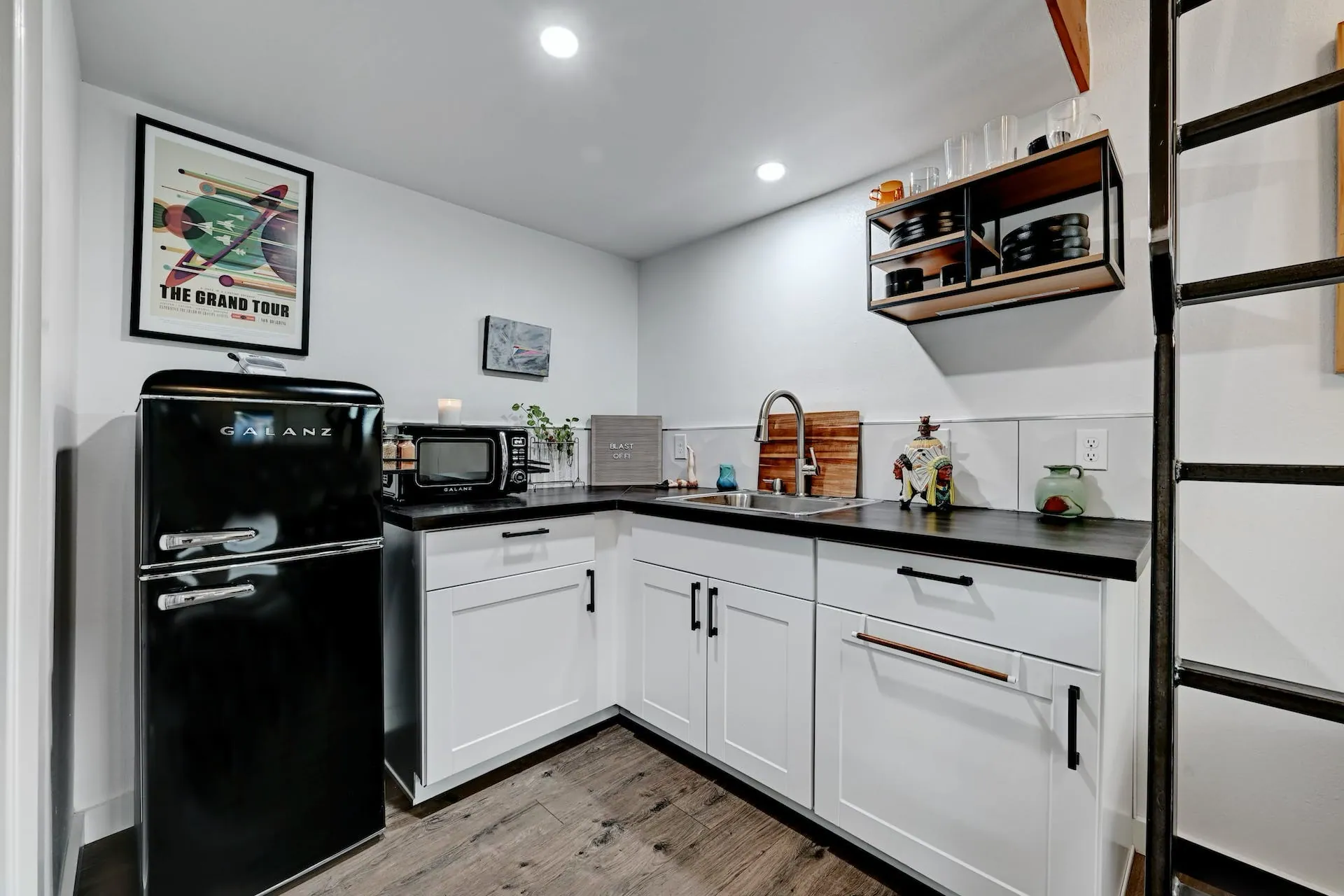 Compact kitchen with black refrigerator, white cabinets, and metal shelving in Domain’s Tuff Shed ADU