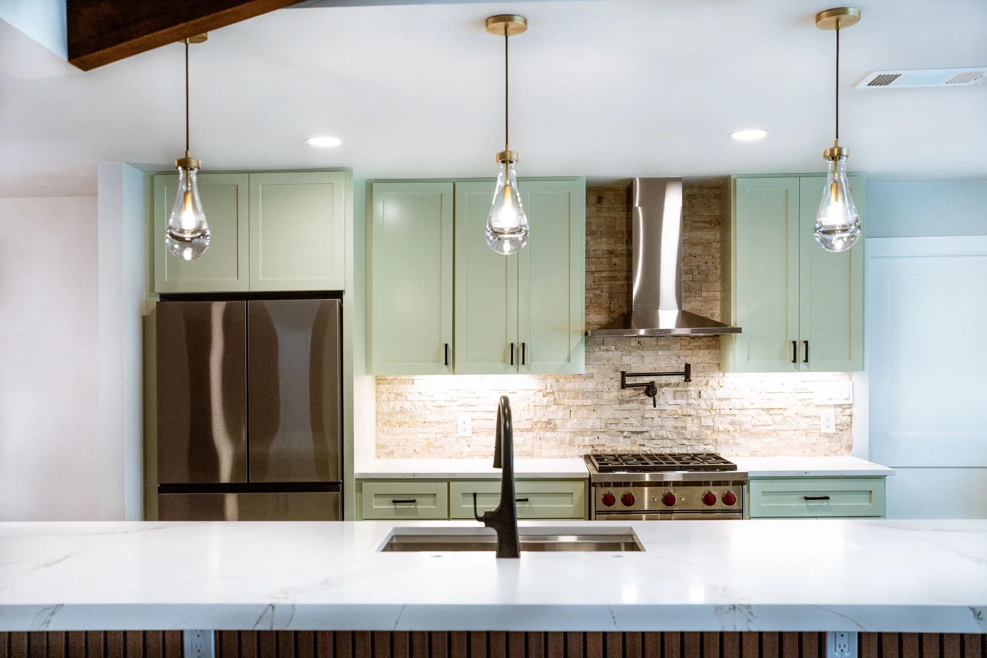 Green cabinetry, stone backsplash, and pendant lights viewed across the island in Domain’s Northwest Austin remodel
